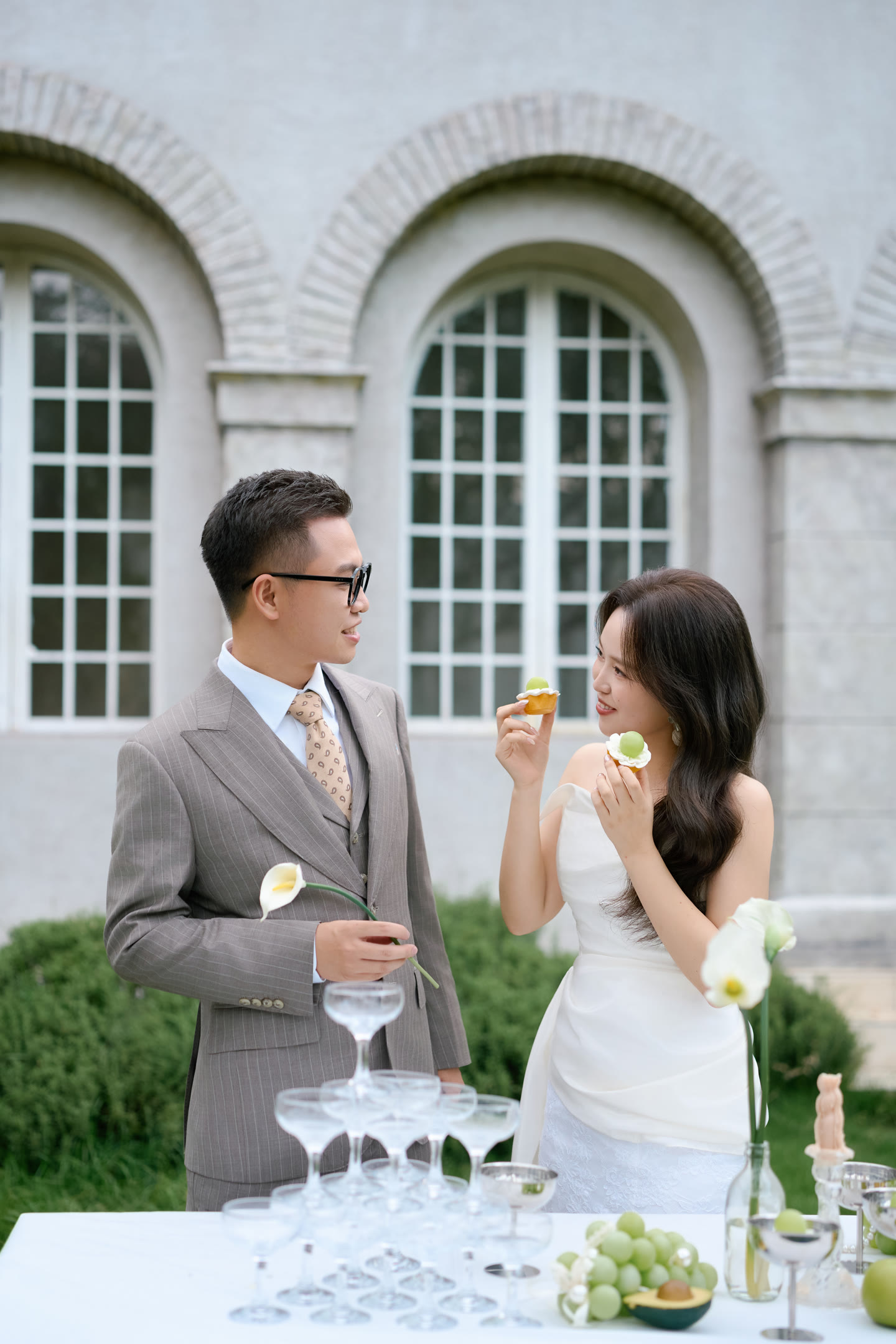宇豪与燕玲在绿色庭院里相视而笑，燕玲手捧葡萄小蛋糕，身旁是白色马蹄莲与香槟杯。